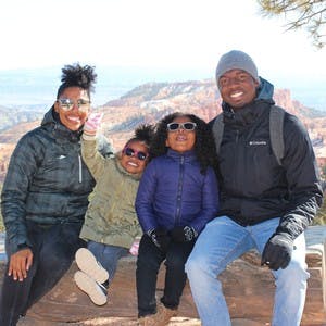 An African American family of 4 wearing winter jackets sits on a rock and smiles for the camera while wearing sunglasses.